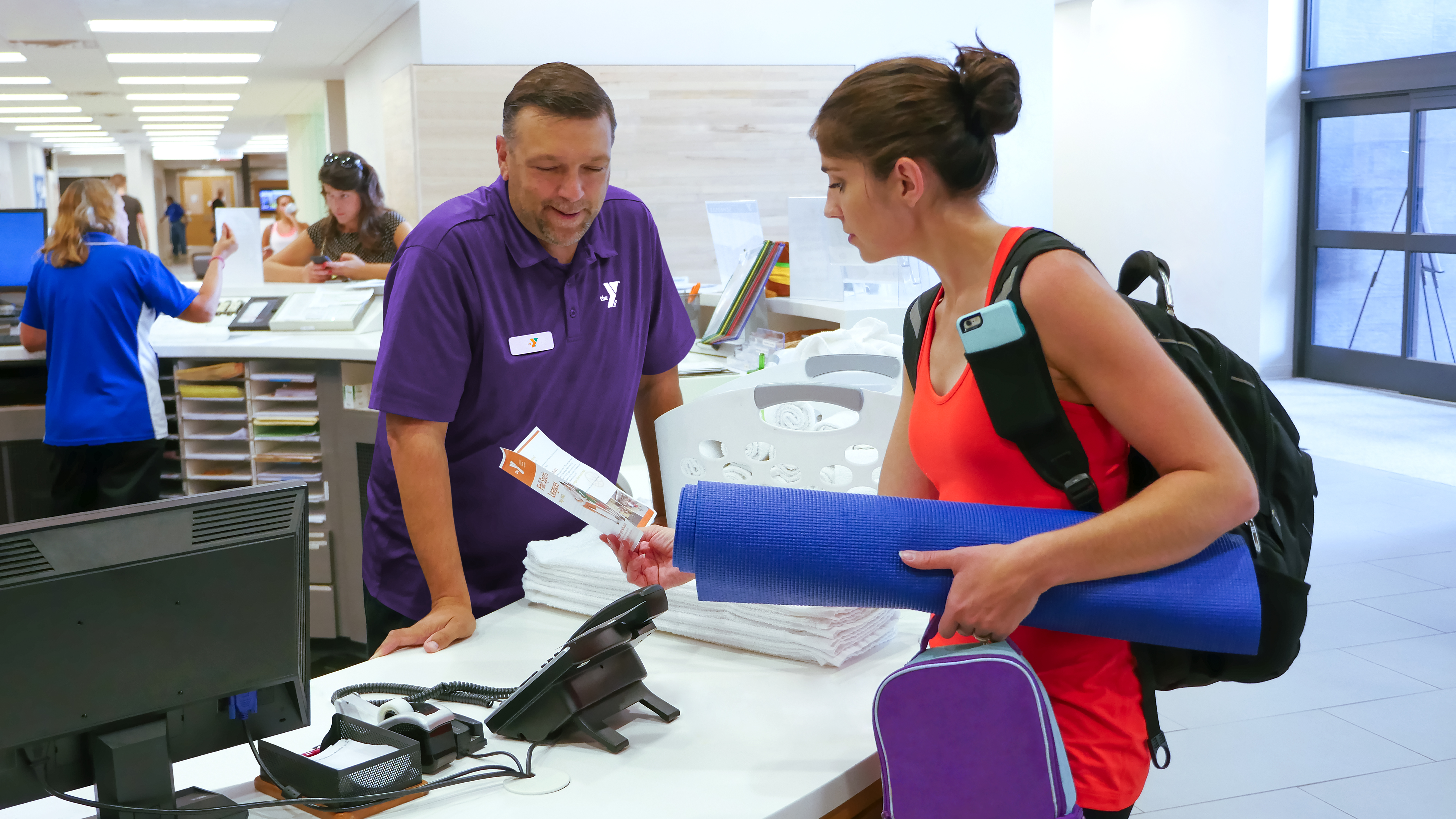 Male Y staff in Y shirt greeting woman holding a yoga mat. 