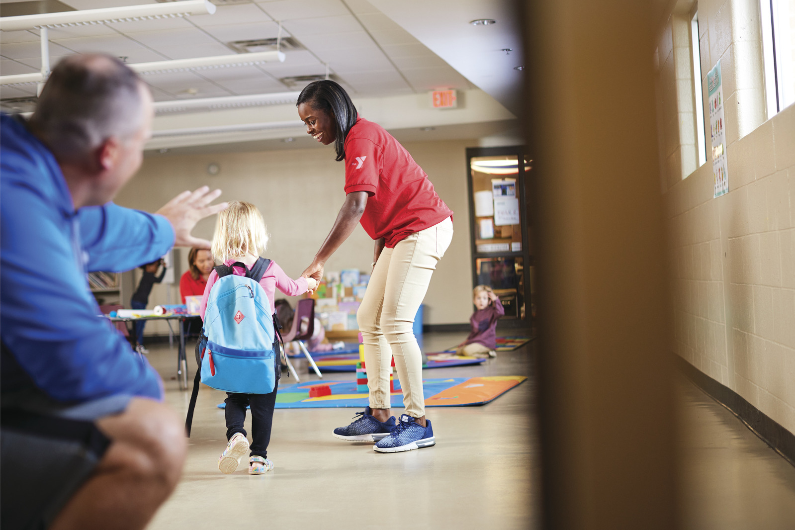 A YMCA staff member helps a child enter a classroom while a parent waves goodbye