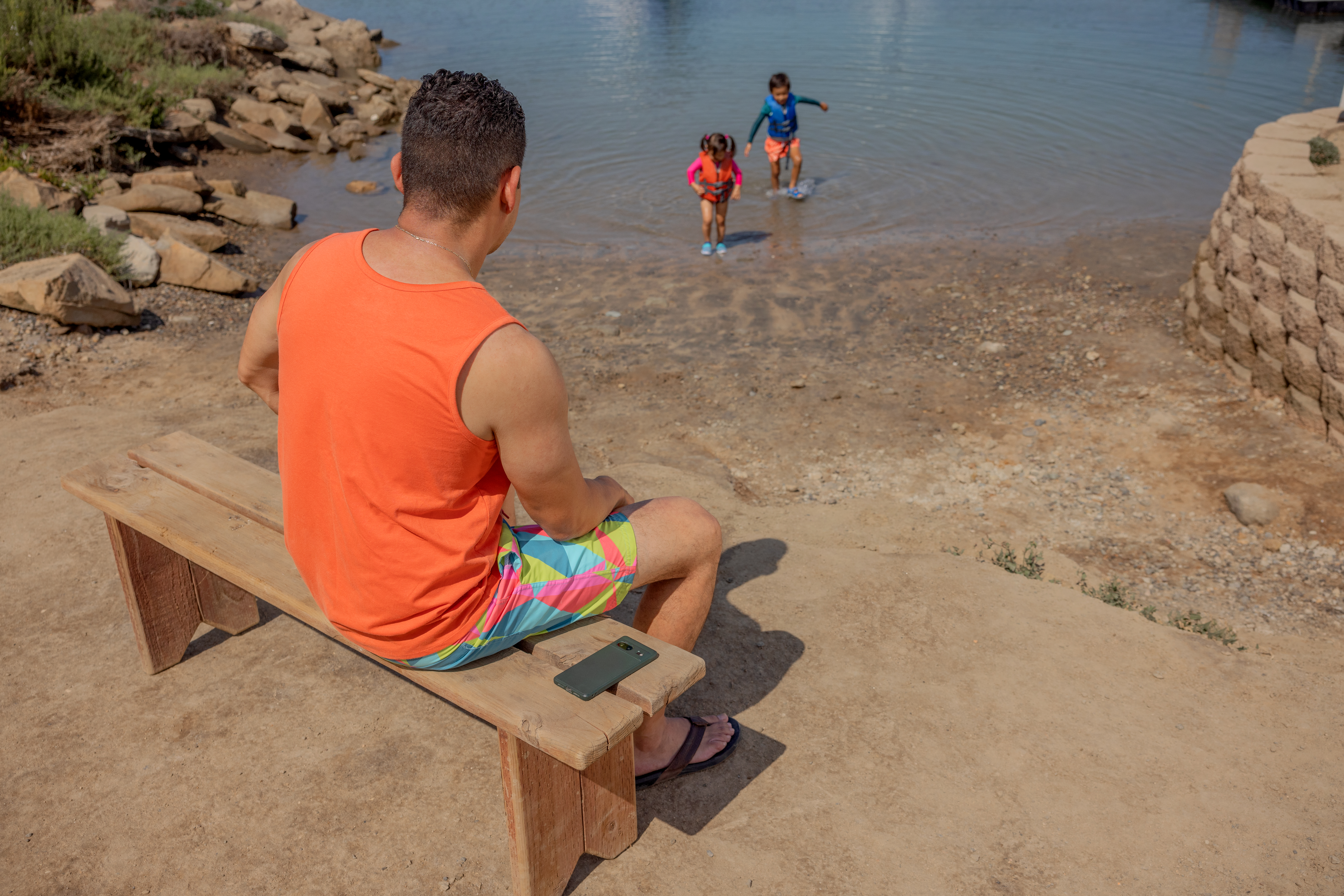 Caregiver watches two youth as they play in the lake, his phone is away from him on the bench